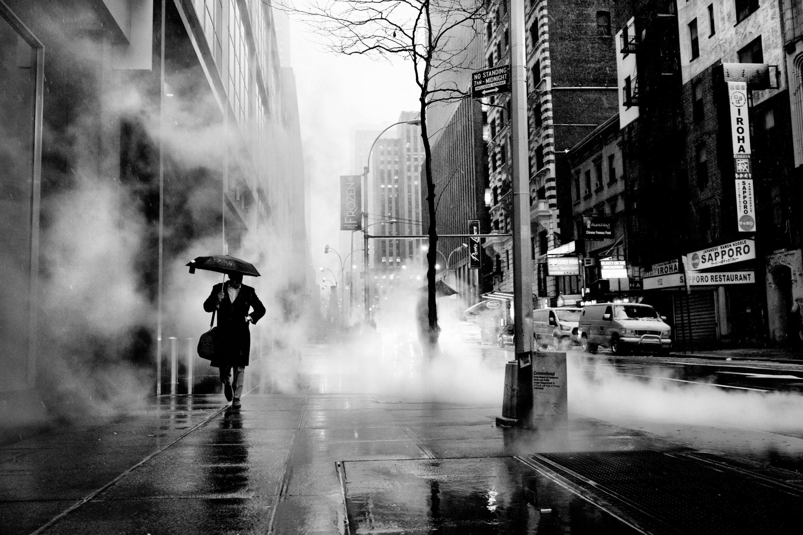 Fotografía urbana en blanco y negro: una persona camina bajo un paraguas entre vapor, edificios y reflejos en la acera